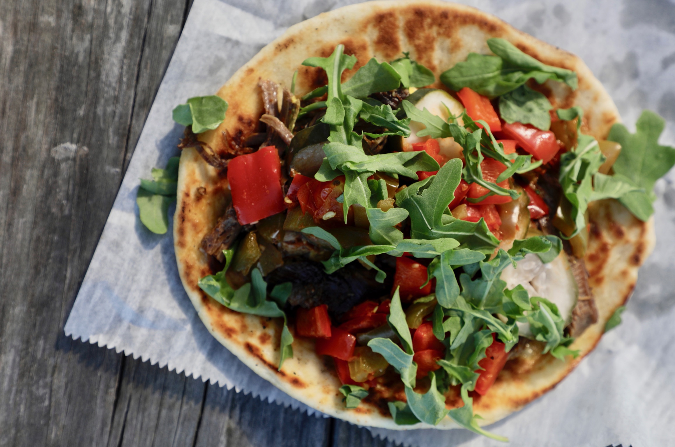 Flatbread piled with arugula and roasted peppers on a weathered table
