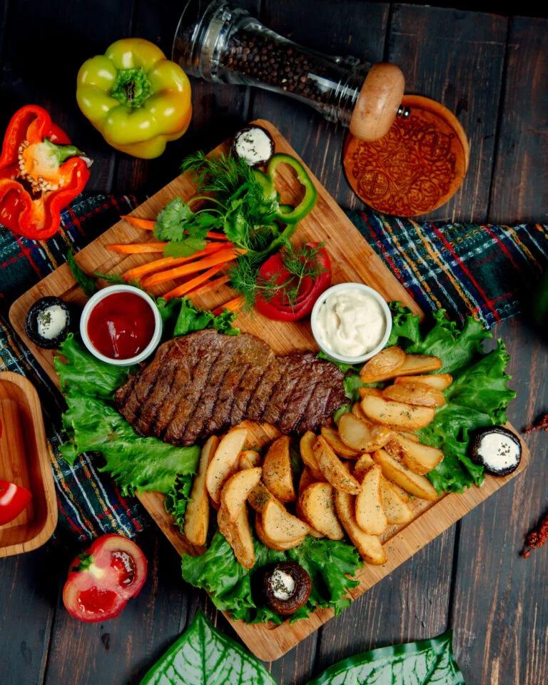 Wooden board with sliced marinated steak, potato wedges, garden vegetables and dipping sauces