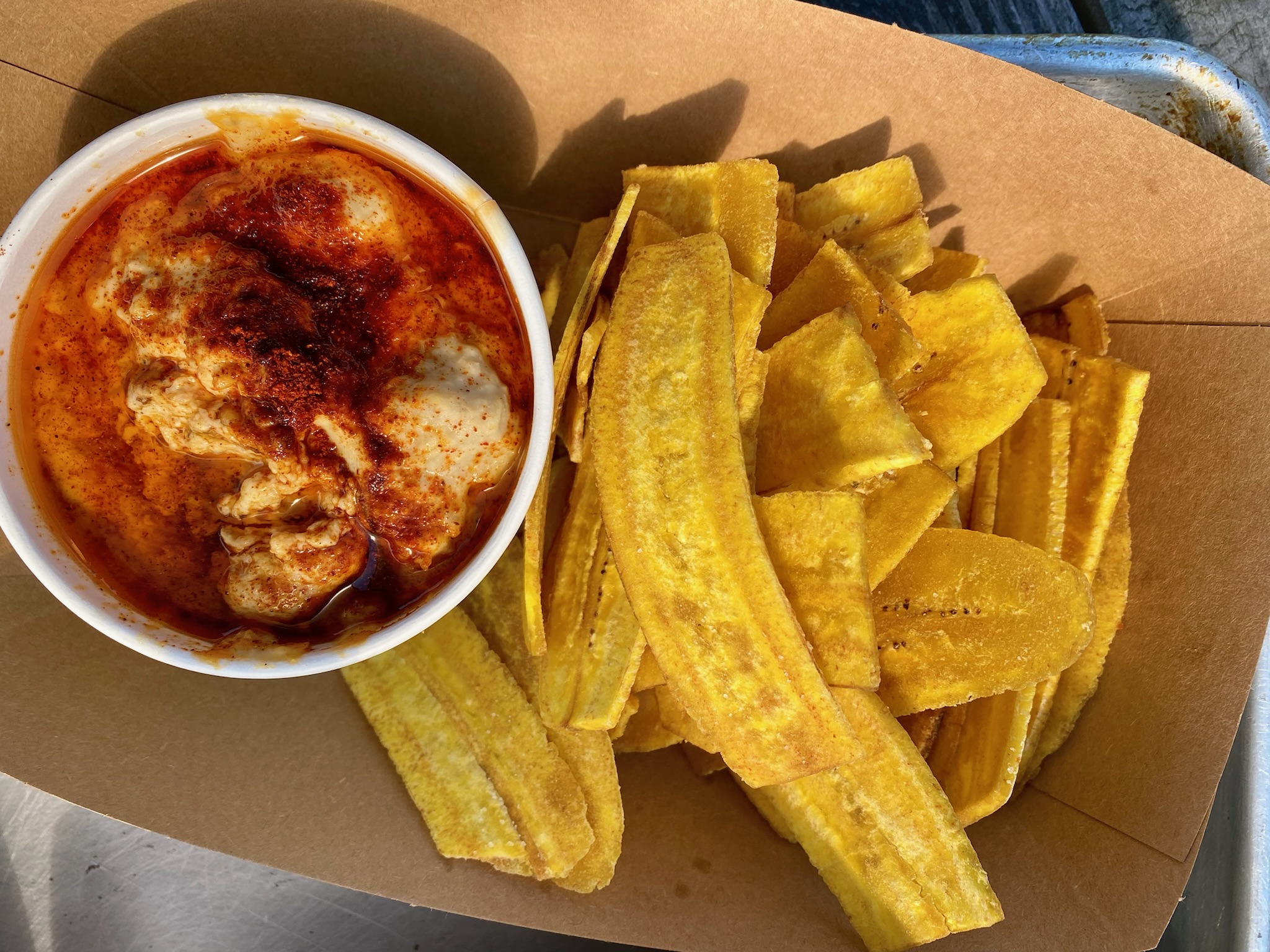 Plantain chips in a paper boat beside a cup of hummus dusted with paprika
