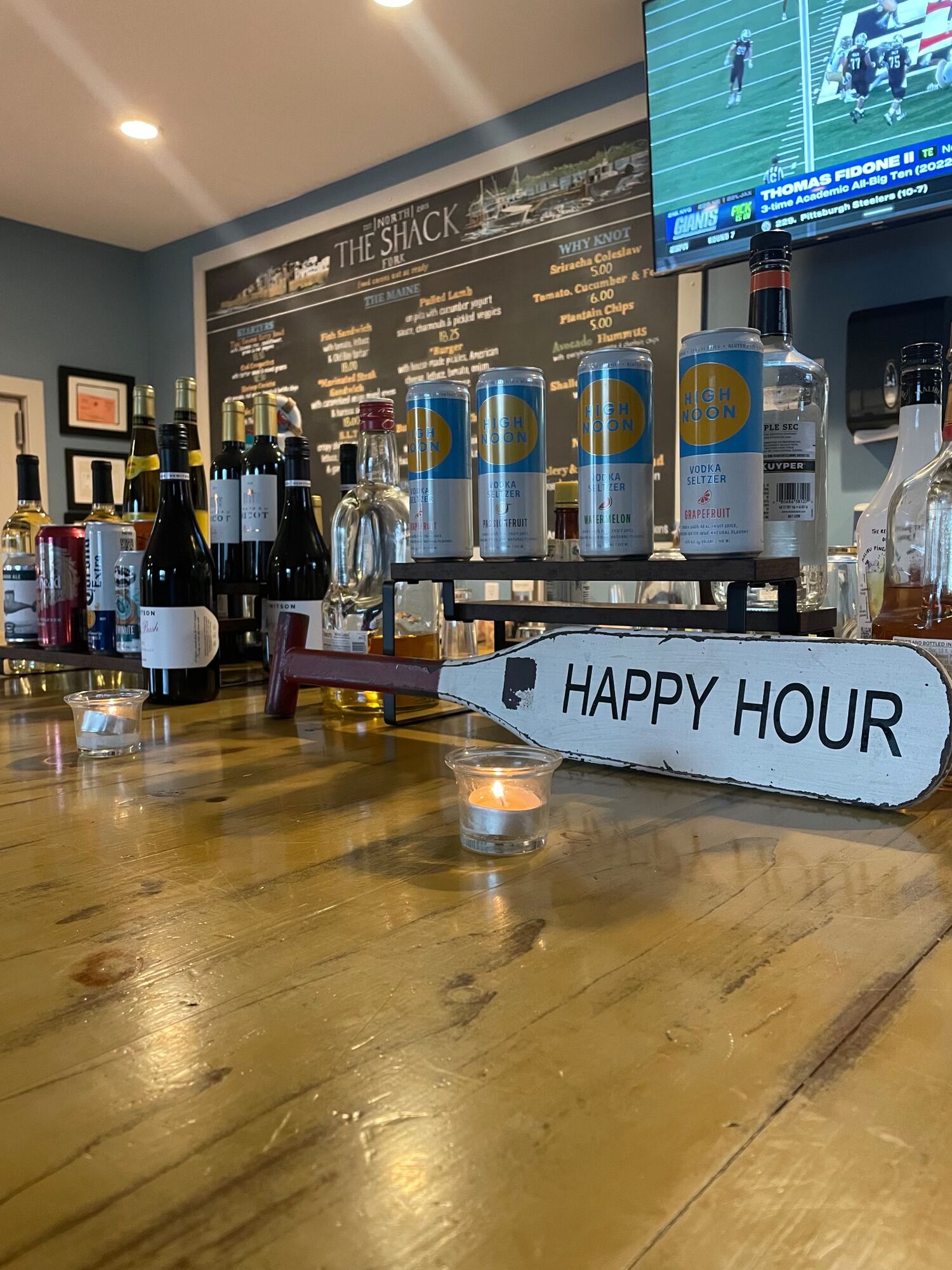 Happy Hour paddle on the wooden bar, with wine, canned seltzers and the chalkboard menu behind