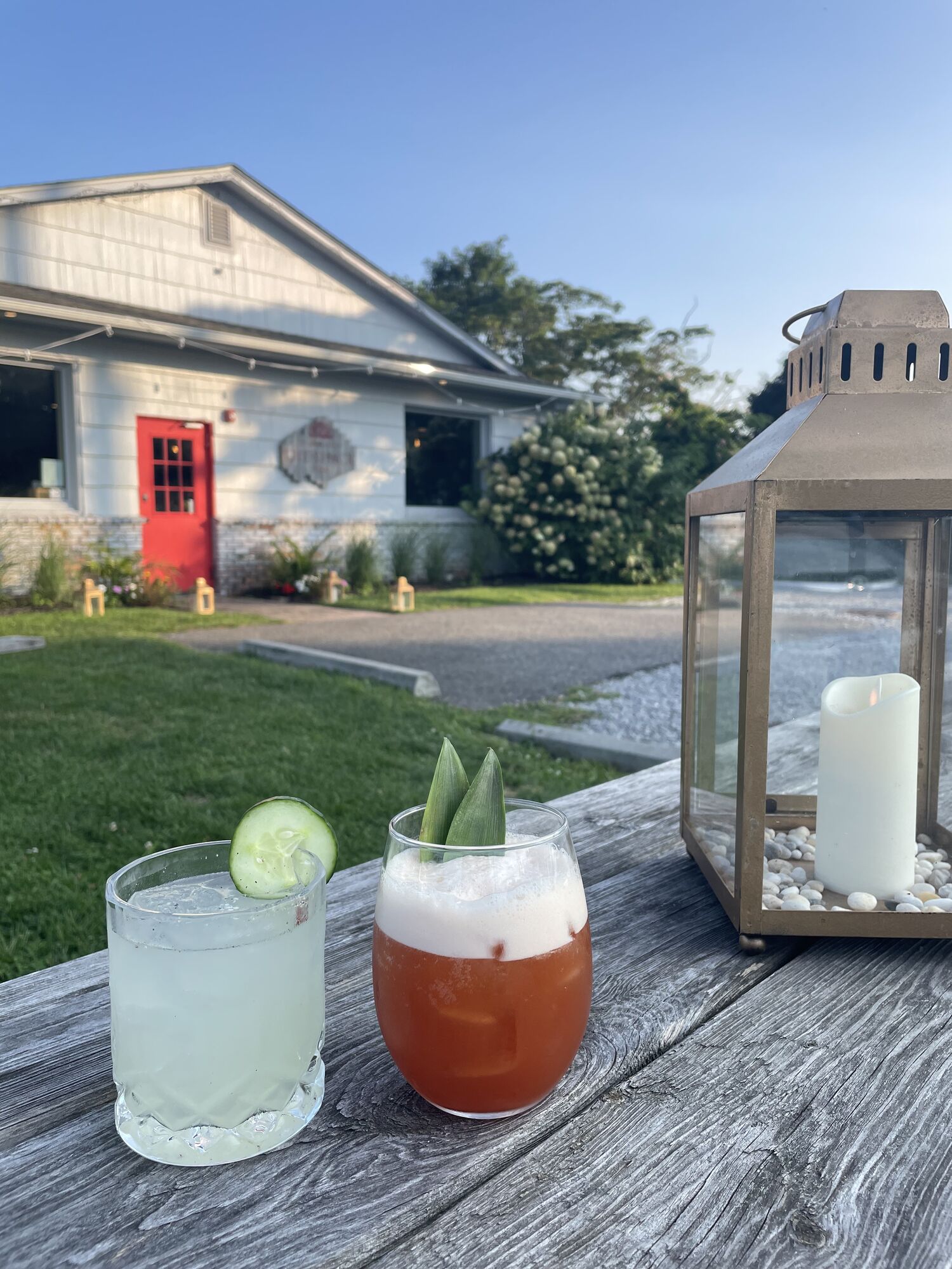 Cucumber cocktail and a foamy red cocktail on a picnic table, with the Shack's red door and a lantern behind