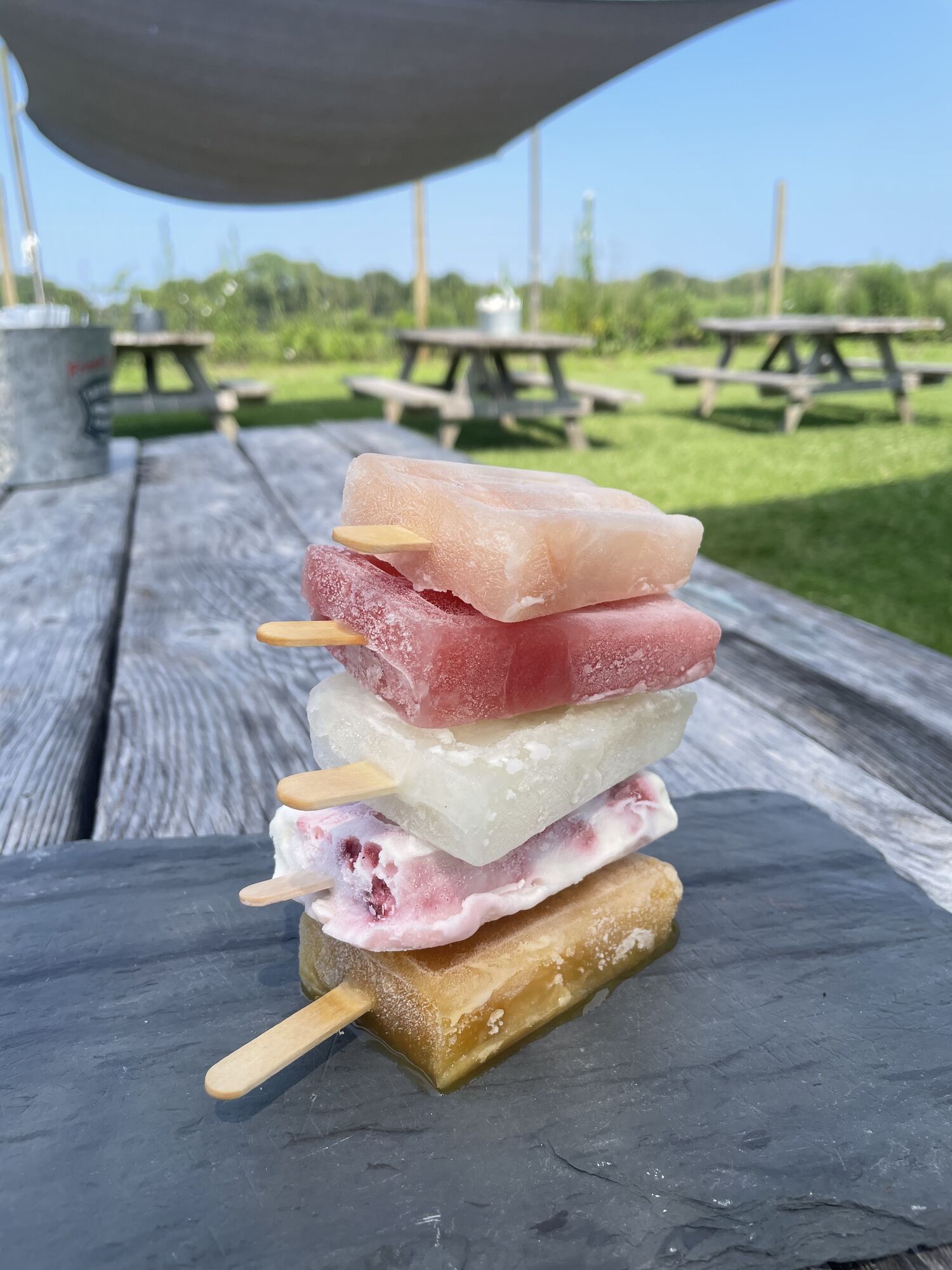 Stack of five house-made fruit popsicles on slate, picnic tables and lawn behind