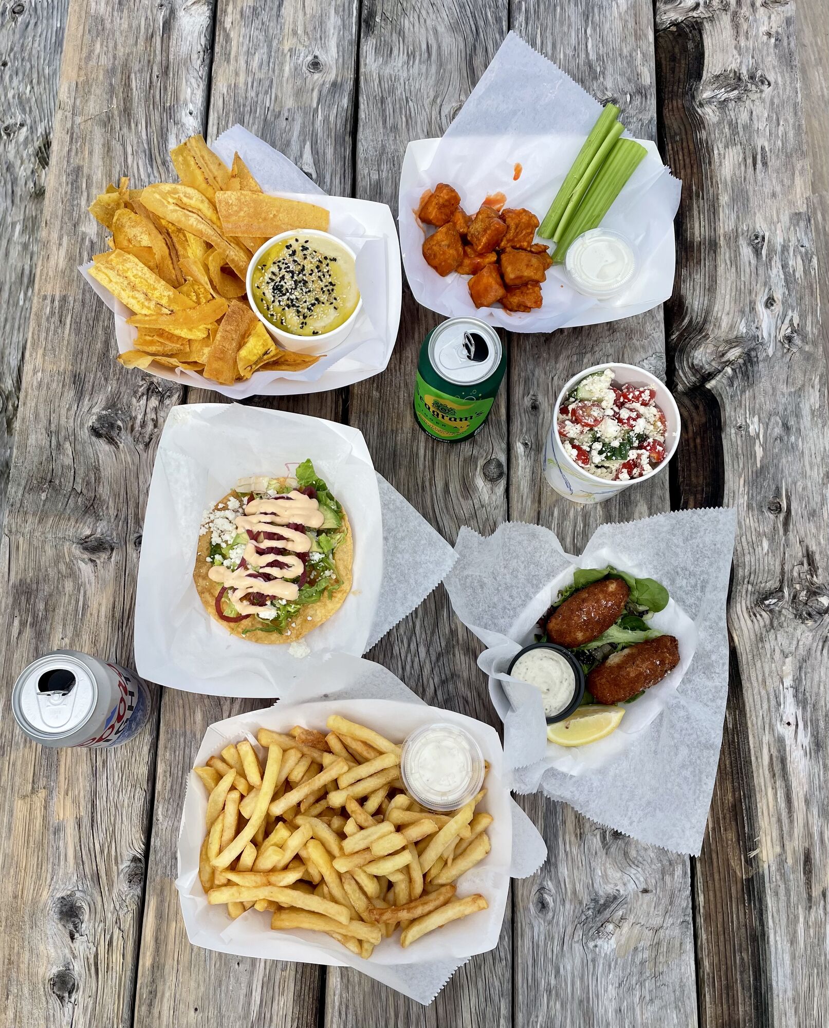 Overhead picnic-table spread of plantain chips with hummus, buffalo cauliflower, a loaded tostada, croquettes, feta salad and fries